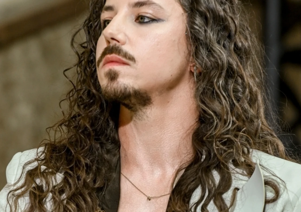 Michał Szpak Michał Szpak w jasnej marynarce z naszyjnikiem, z charakterystycznymi długimi włosami i makijażem oczu.