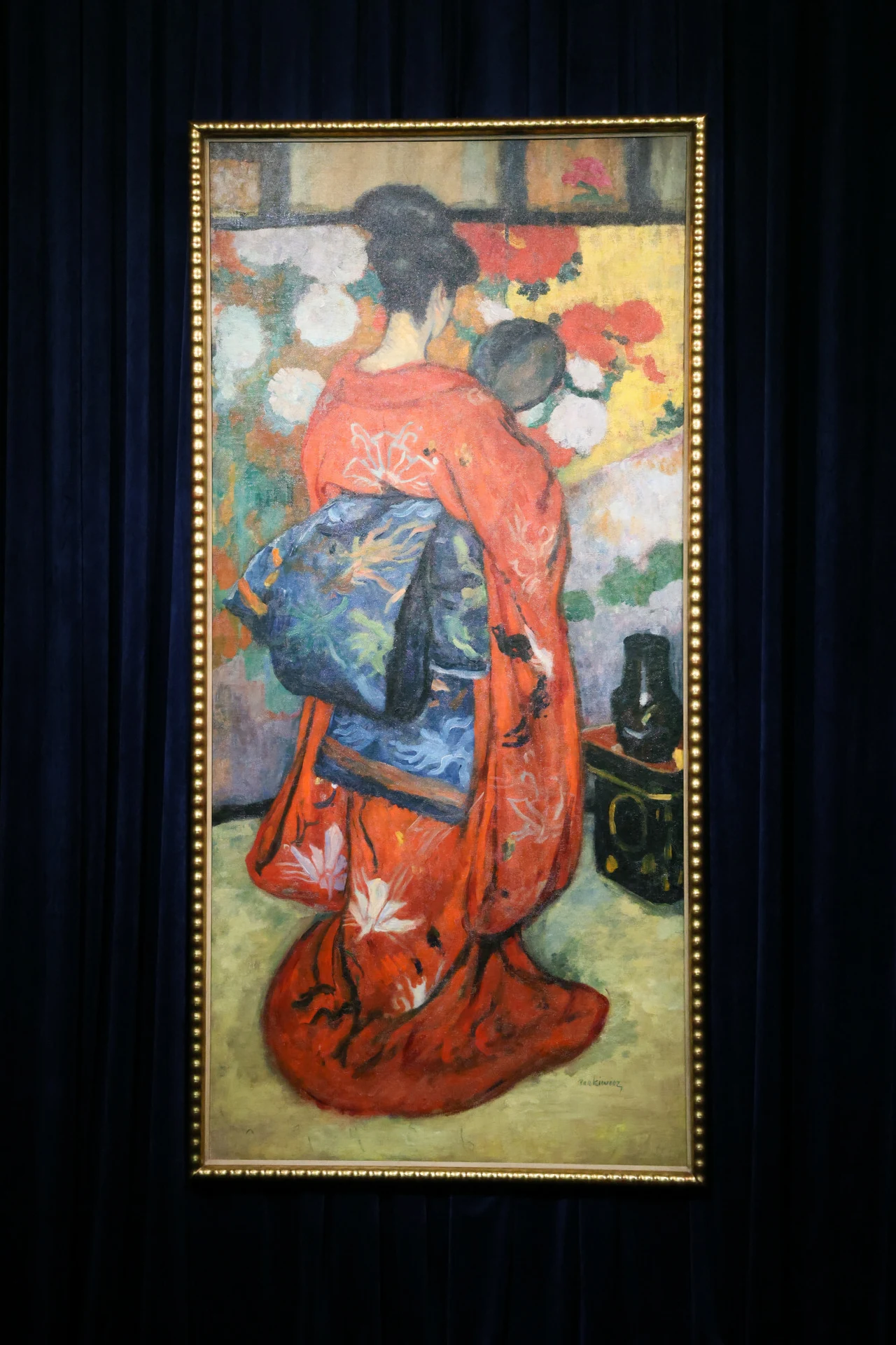 Kobieta ubrana w tradycyjne japońskie kimono z dzieckiem na rękach, zwrócone tyłem do widza, na tle kolorowej, kwiecistej dekoracji.