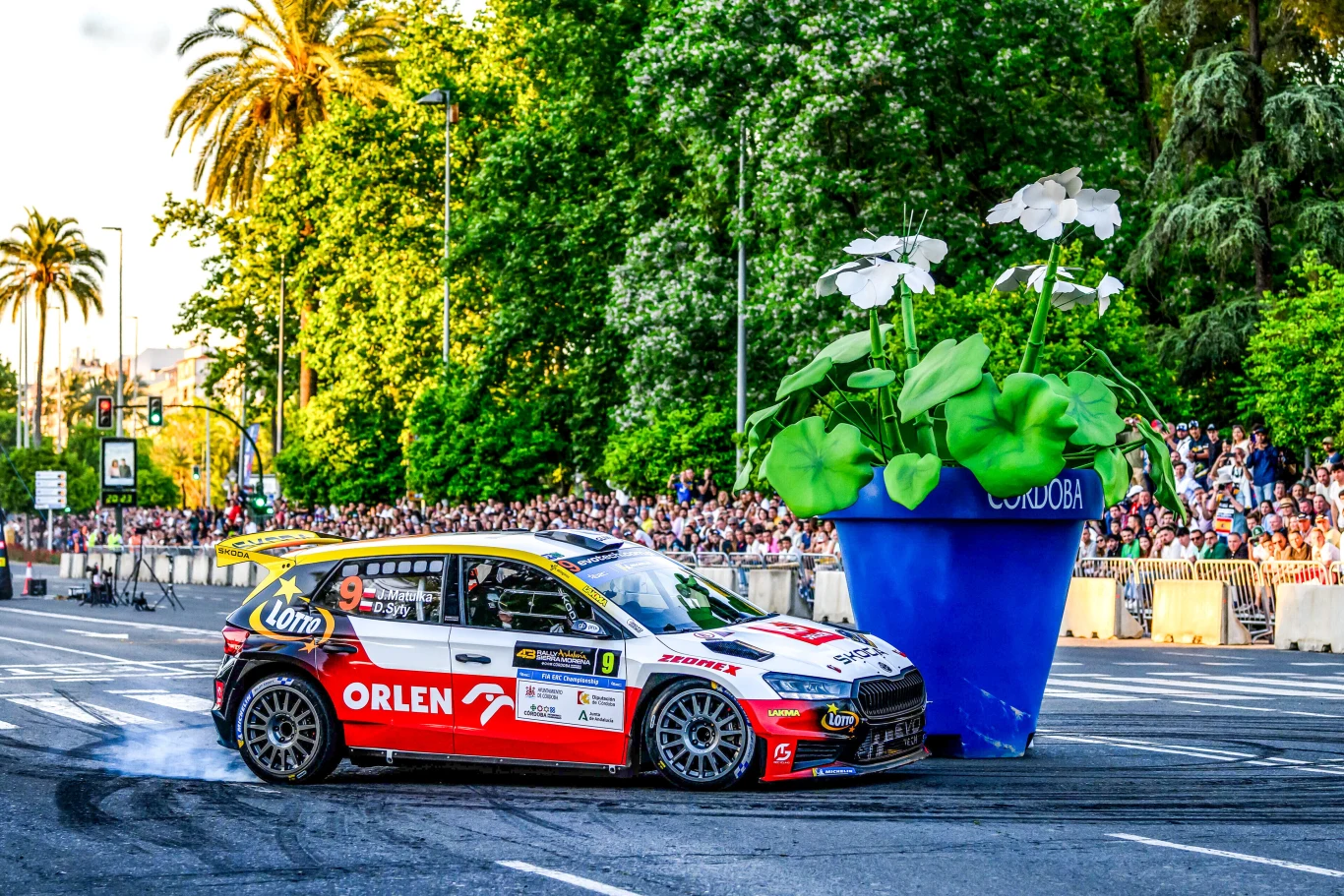 Jakub Matulka i Damian Syty w tym roku bronią tytułu mistrzów Polski Sportowy samochód rajdowy w barwach Orlen wykonujący efektowny drift na asfaltowej nawierzchni, tuż obok dużej, niebieskiej donicy z białymi kwiatami; w tle tłum kibiców i zieleń drzew.