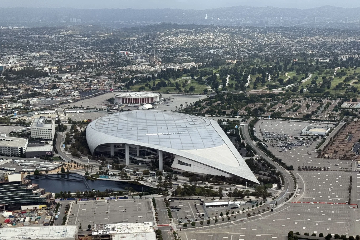 SoFi Stadium wLos Angeles. Piłkarskie MŚ 2026 odbędą się na najnowocześniejszych obiektach sportowych. Nowoczesny stadion o futurystycznej konstrukcji otoczony rozległymi parkingami i terenami miejskimi, w tle niskie zabudowania i zielone pola, a dalej panorama miasta sięgająca po horyzont.