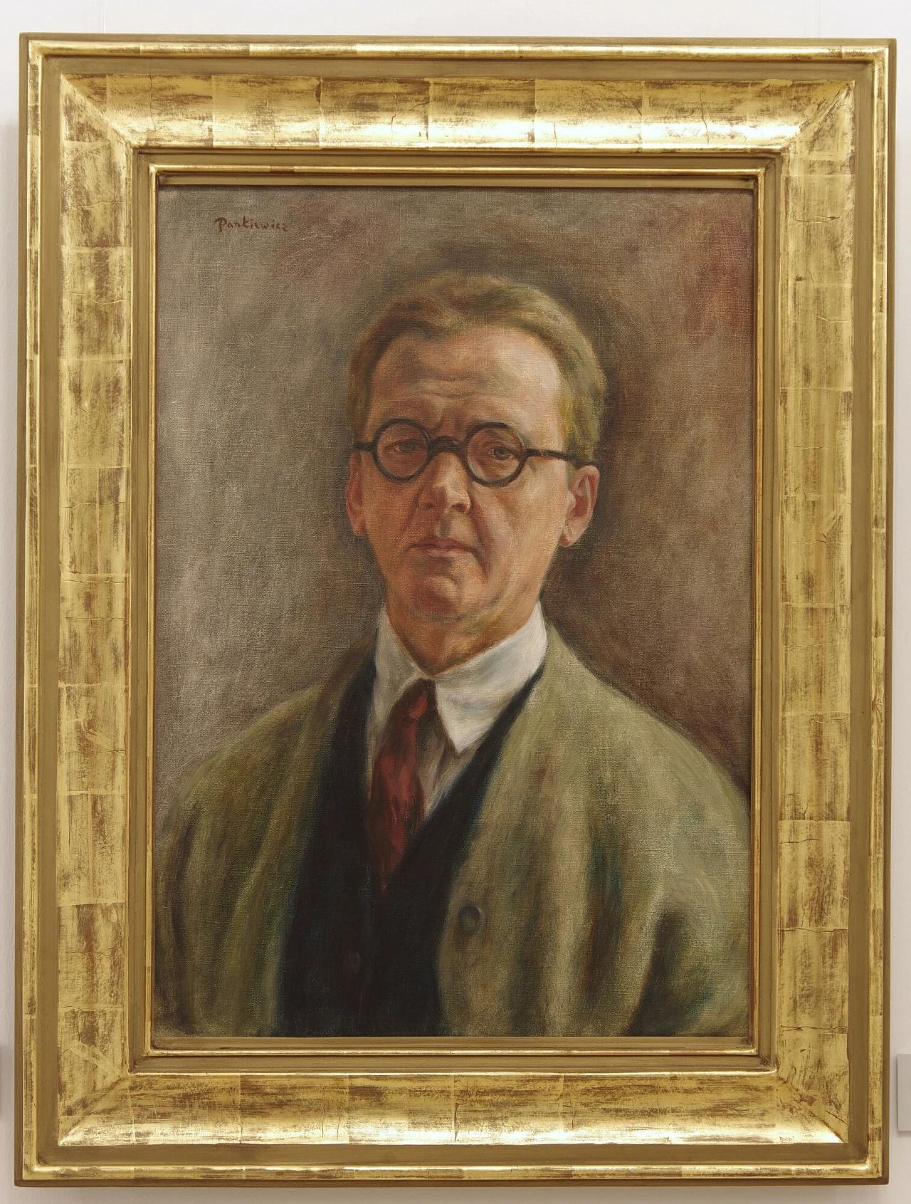 Portret mężczyzny w okularach z krótkimi, jasnobrązowymi włosami, ubrany w jasną koszulę, ciemny krawat oraz zielonkawy płaszcz, umieszczony w ozdobnej, złotej ramie.