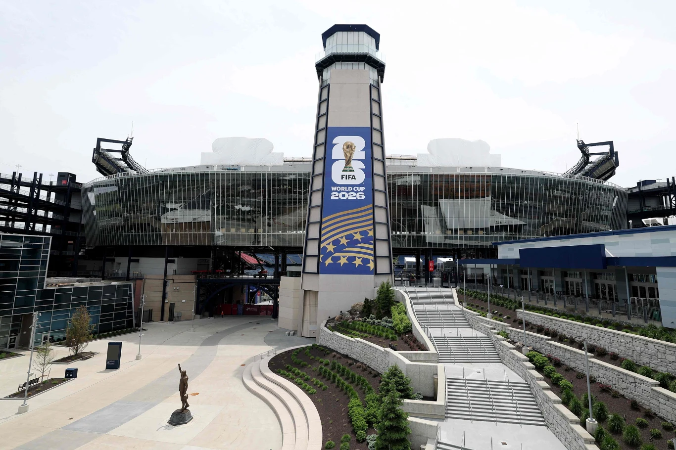 Gillette Stadium w Foxborough pomieści około 66 tys. widzów. Nowoczesny stadion z dużym banerem FIFA World Cup 2026, szerokimi schodami prowadzącymi do wejścia i rzeźbą stojącą przy starannie zagospodarowanej zieleni.