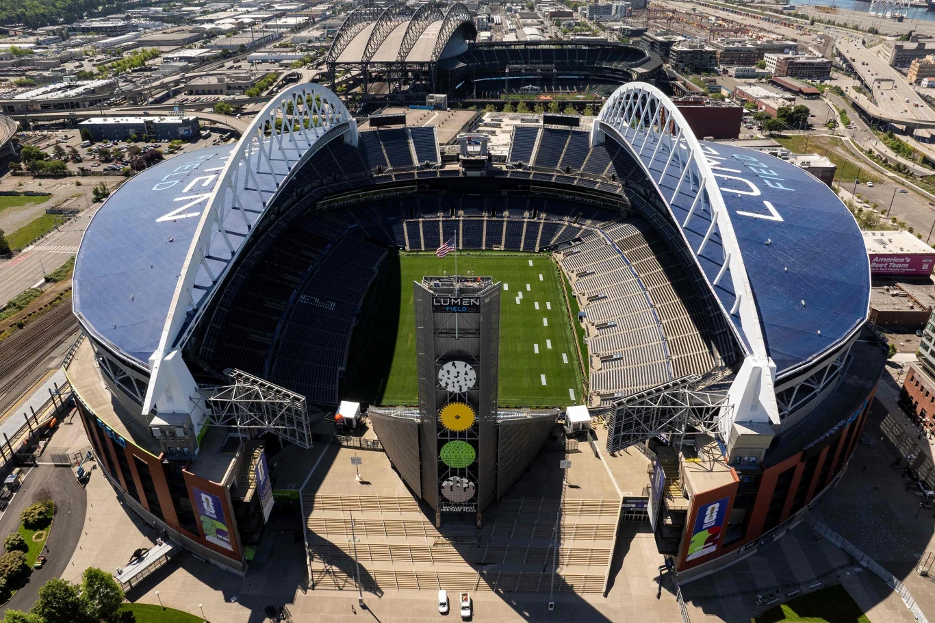 Stadion Lumen Field w Seattle ma wyjątkowy układ trybun. Duży, nowoczesny stadion z charakterystycznymi łukowatymi zadaszeniami, zieloną murawą pośrodku i trybunami otaczającymi całą arenę sportową.