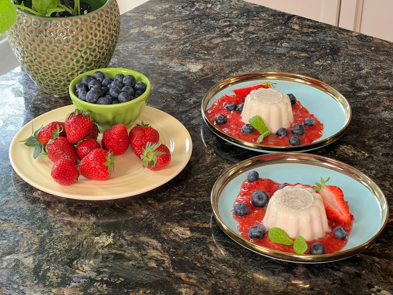 Deser rodem z Włoch, panna cotta - przepis Ewy Wachowicz Dwie porcje deseru panna cotta udekorowane borówkami, truskawkami i listkami mięty na kolorowych talerzach, obok talerz z truskawkami oraz miseczka z borówkami na marmurowym blacie.