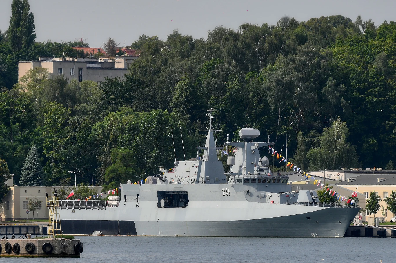 nowoczesna fregata marynarki wojennej zacumowana przy nabrzeżu, ozdobiona flagami sygnałowymi, na tle zieleni i budynków