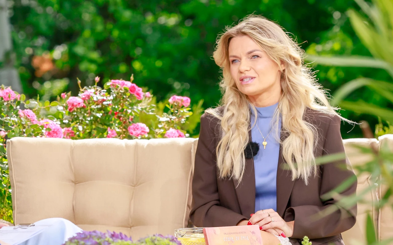 Emilia Komarnicka w "halo tu polsat" Kobieta z długimi blond włosami siedząca na beżowej sofie w ogrodzie, otoczona kwitnącymi różowymi kwiatami, ubrana w brązową marynarkę i niebieską bluzkę, z mikrofonem przypiętym do ubrania.