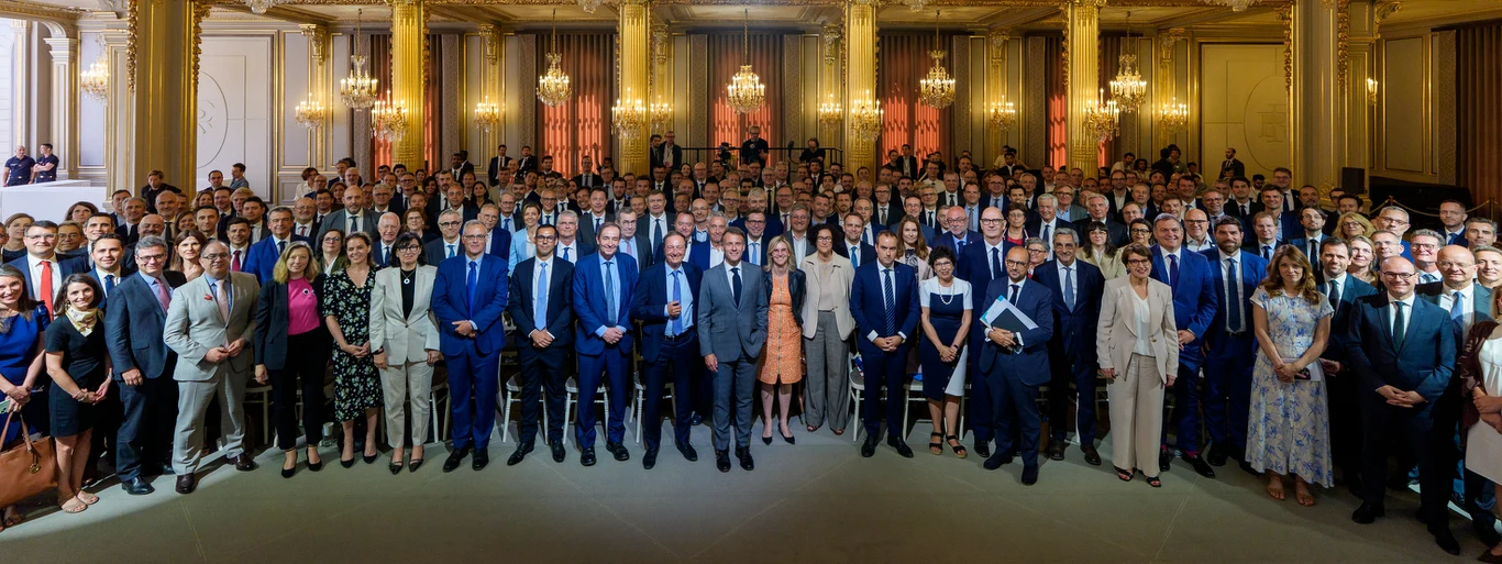 Duża grupa elegancko ubranych osób zgromadzona w reprezentacyjnej sali z wysokimi sufitami, złotymi kolumnami i kryształowymi żyrandolami.