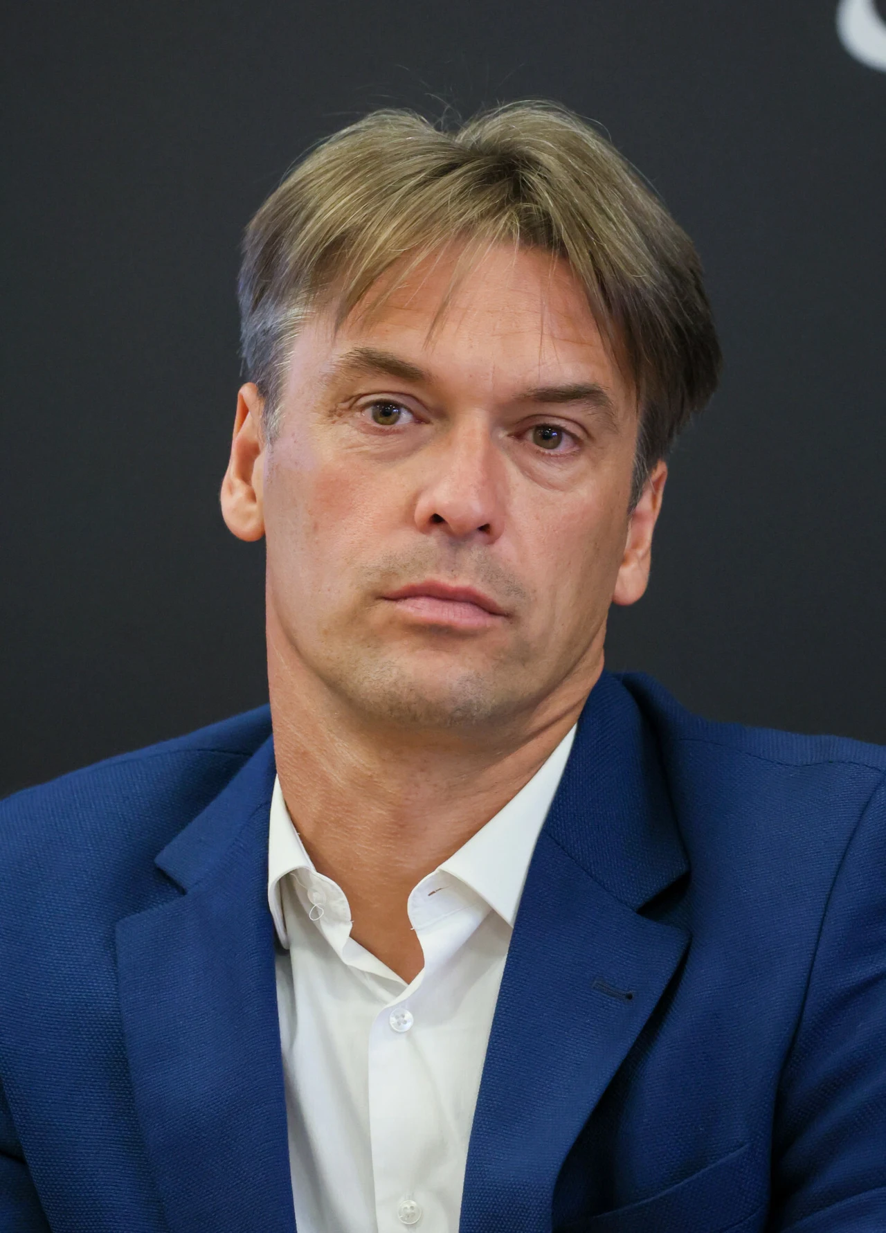 Mężczyzna w średnim wieku o jasnych włosach i poważnym wyrazie twarzy, ubrany w granatową marynarkę i białą koszulę, siedzi na tle ciemnego, jednolitego tła.