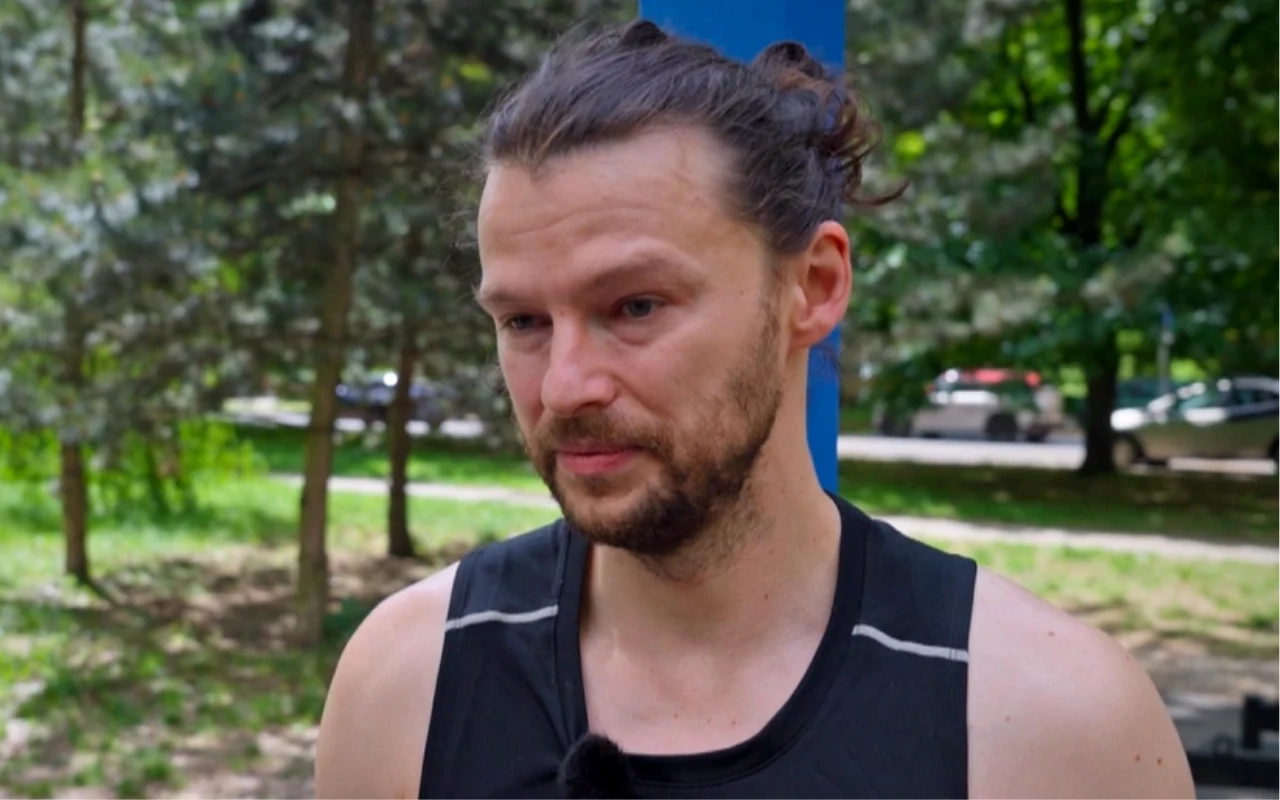 Mateusz Kościukiewicz Mężczyzna z brodą i włosami związanymi w kucyk, ubrany w czarną sportową koszulkę, stoi na tle zielonych drzew i parku.