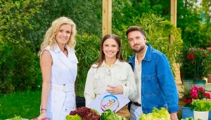 Trzy uśmiechnięte osoby stoją za stołem przykrytym niebiesko-białym obrusem, na którym ułożone są różne gatunki sałat i ziół oznaczone kartonikami z nazwami, a w tle widoczne są rośliny doniczkowe i drewniana altana.