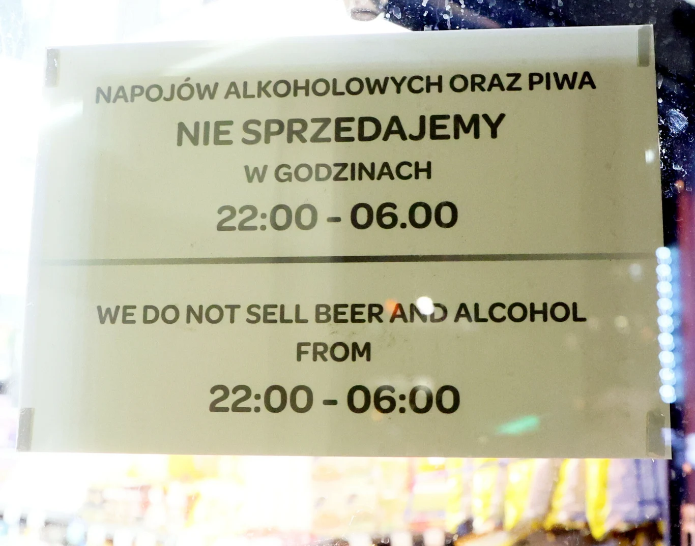 Biała tablica z czarnym napisem informującym w języku polskim i angielskim o zakazie sprzedaży alkoholu i piwa w godzinach 22:00–06:00, umieszczona za szklaną szybą.