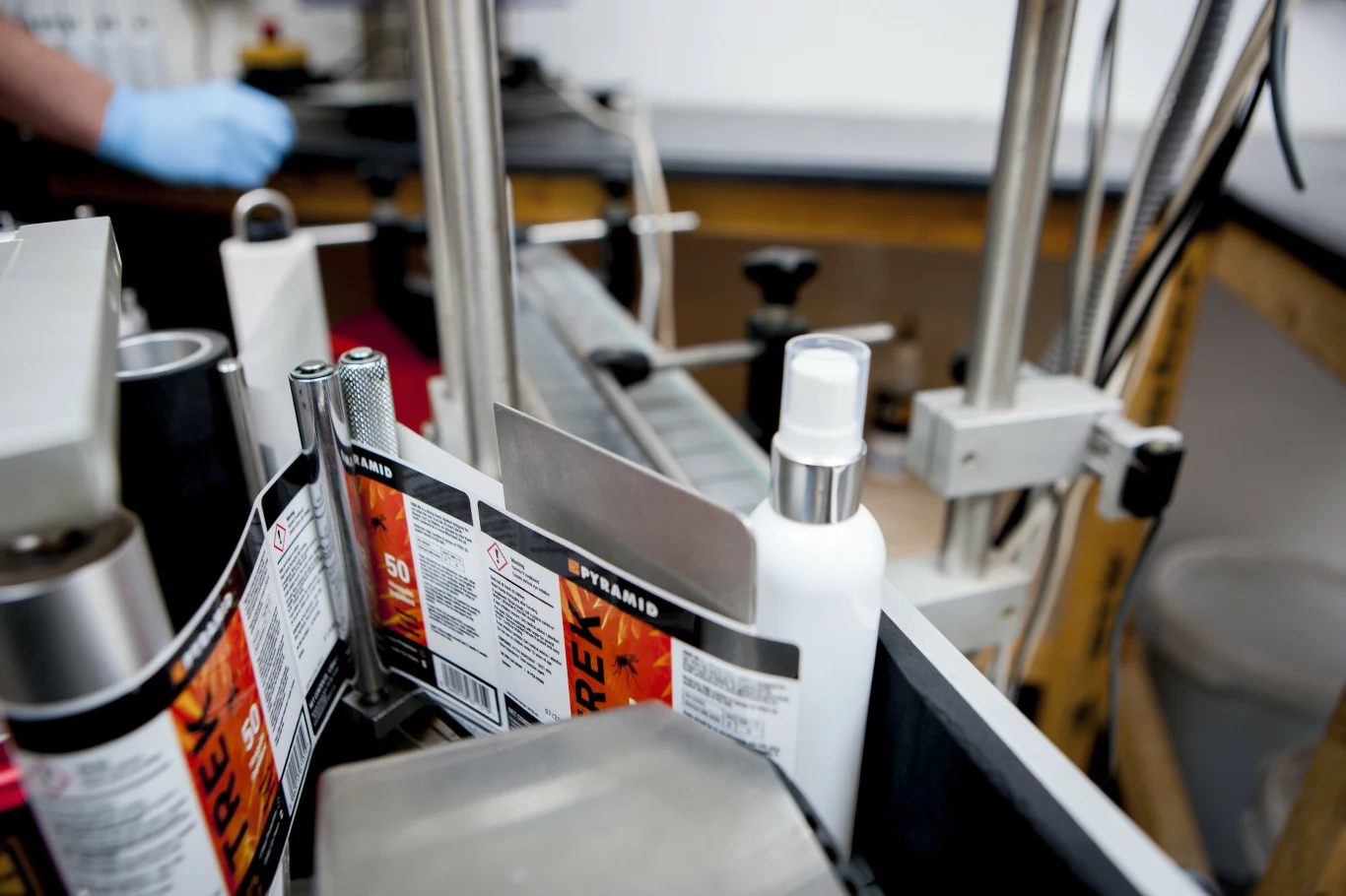 Biała butelka ustawiona pomiędzy maszynami do etykietowania, obok widoczne rolki z kolorowymi etykietami oraz elementy stalowej konstrukcji linii produkcyjnej.