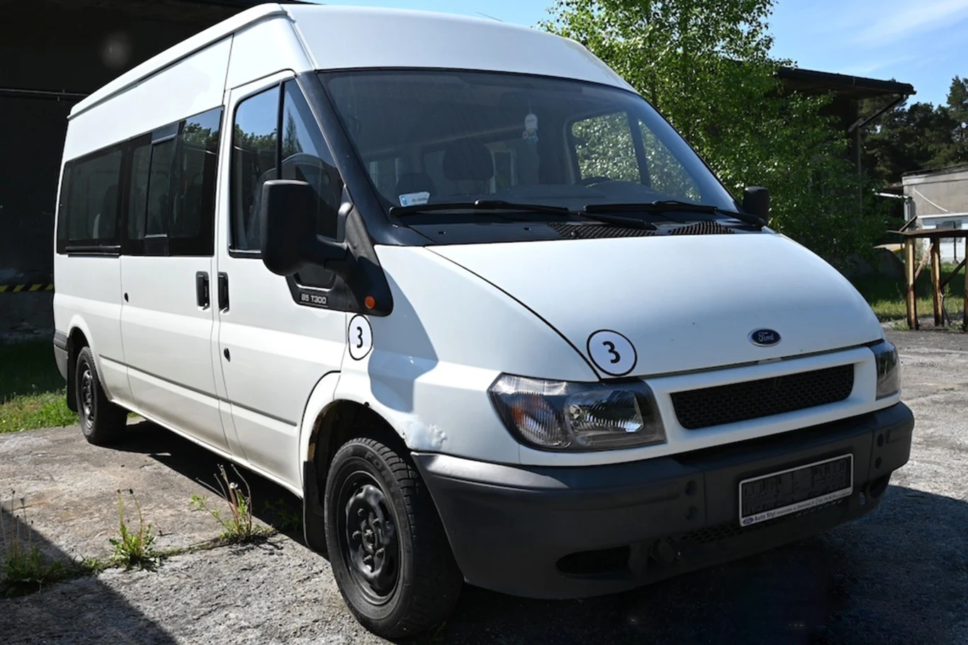 Biały bus Ford Transit z czarnymi zderzakami i oznaczeniami liczby 3 na masce oraz błotniku, zaparkowany na betonowej nawierzchni obok zieleni.