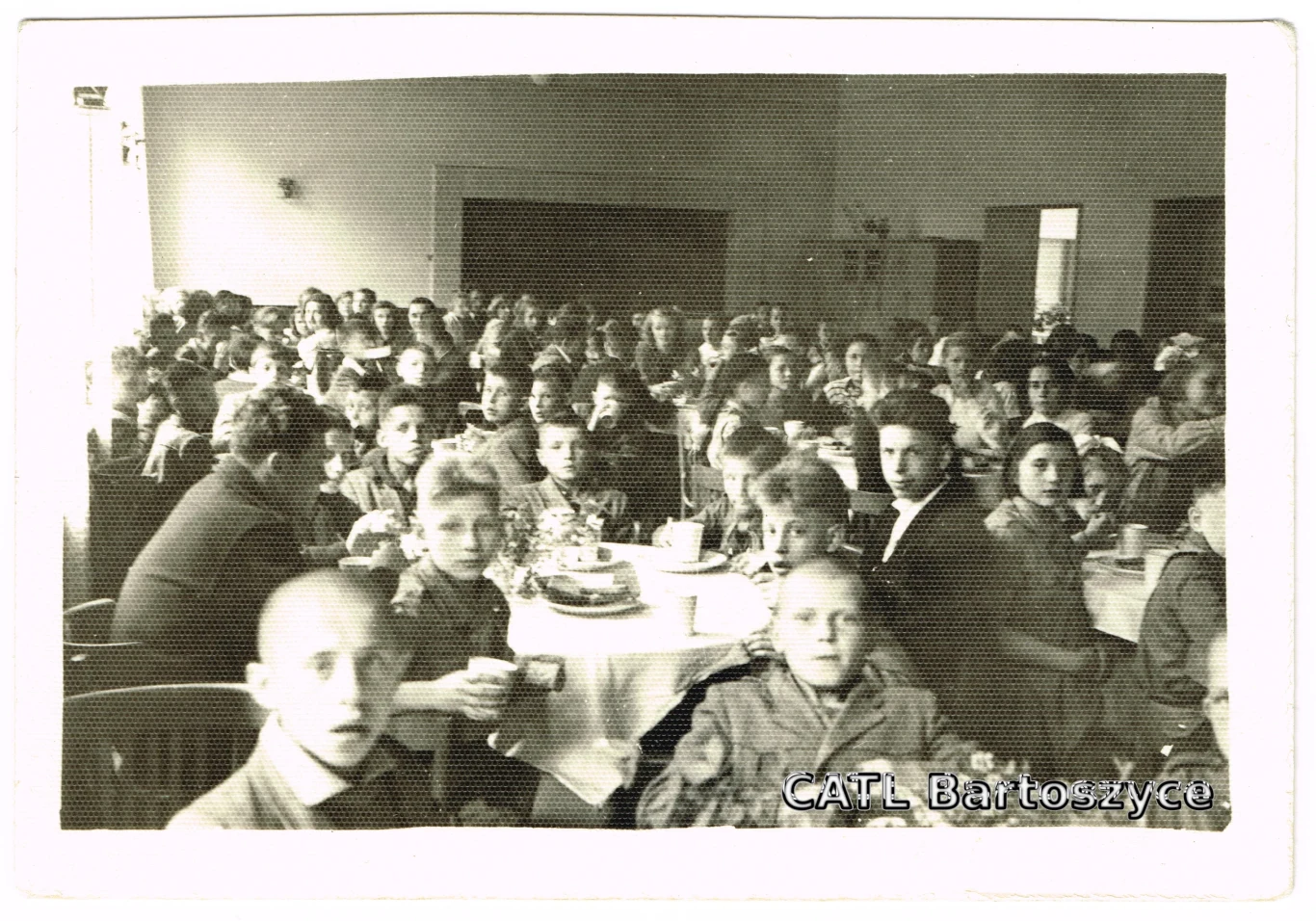 Dzieci z Domu Dziecka przy śniadaniu, Bartoszyce, 1952 Duża grupa dzieci i dorosłych siedzących przy stołach zastawionych ciastem i napojami, skupionych na wspólnym wydarzeniu w przestronnym, jasnym pomieszczeniu.