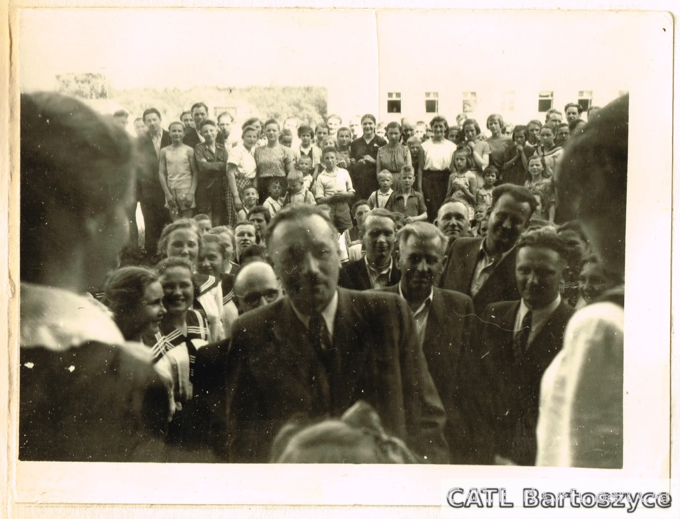 Wizyta Bolesława Bieruta w bartoszyckim Domu Dziecka, 1953 Duża grupa ludzi ustawionych w kilku rzędach, z przodu dorosłe osoby w garniturach i kobiety, w głębi dzieci i młodzież, wszyscy uśmiechnięci i skierowani w stronę aparatu, w tle fragmenty budynku i drzewa.