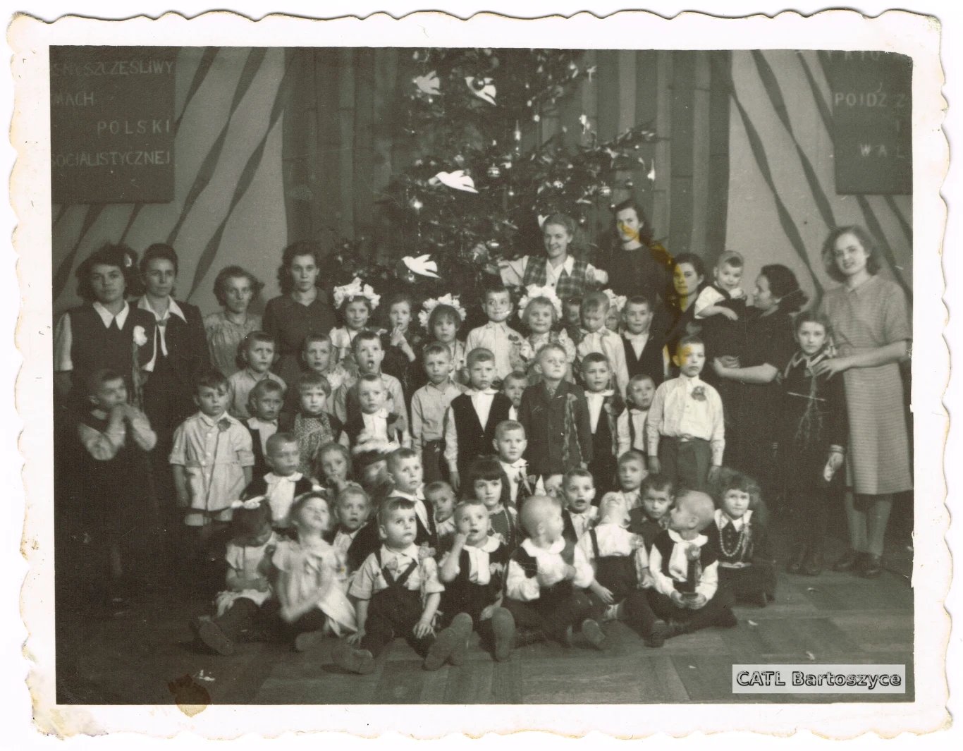 Zdjęcie grupowe na tle choinki, Bartoszyce, 1949 Grupa dzieci i dorosłych ubranych odświętnie ustawiona przed dużą choinką ozdobioną papierowymi dekoracjami, całość na tle poziomych pasów i tablic z napisami.