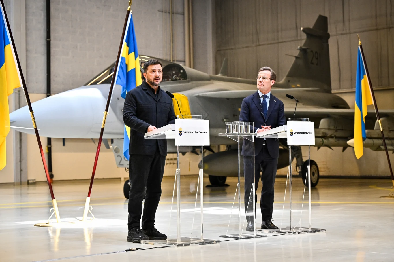 Podczas wizyty Wołodymyra Zełenskiego w szwedzkiej bazie lotniczej Ärna, ogłoszono, że Ukraina otrzyma nieodpłatnie 16 myśliwców JAS-39C/D Gripen. Przy tym Ukraina zakupiła 20 Gripenów E za 2,5 mld euro, które zostaną sfinansowane z pakietu wsparcia finansowego dla Ukrainy zatwierdzonego przez Komisję Europejską. Dwóch mężczyzn stoi przy przezroczystych mównicach ustawionych na tle myśliwca wojskowego oraz dwóch flag Ukrainy i dwóch flag Szwecji.