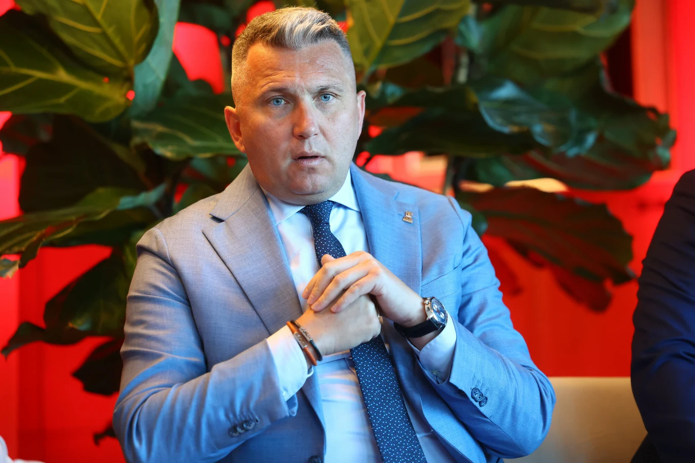 Mężczyzna w jasnoniebieskim garniturze siedzi z dłońmi splecionymi przed sobą, w tle duże zielone liście roślin na tle czerwonej ściany.