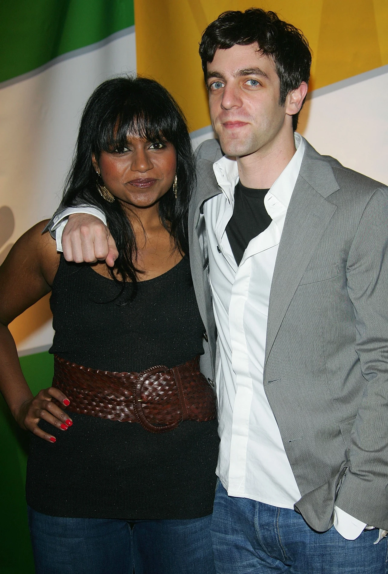 Mindy Kaling i B.J. Novak, 2006 r. Dwie osoby stojące obok siebie, kobieta z długimi ciemnymi włosami w czarnej bluzce i szerokim brązowym pasku oraz mężczyzna w białej koszuli i szarej marynarce, na tle kolorowej ściany.