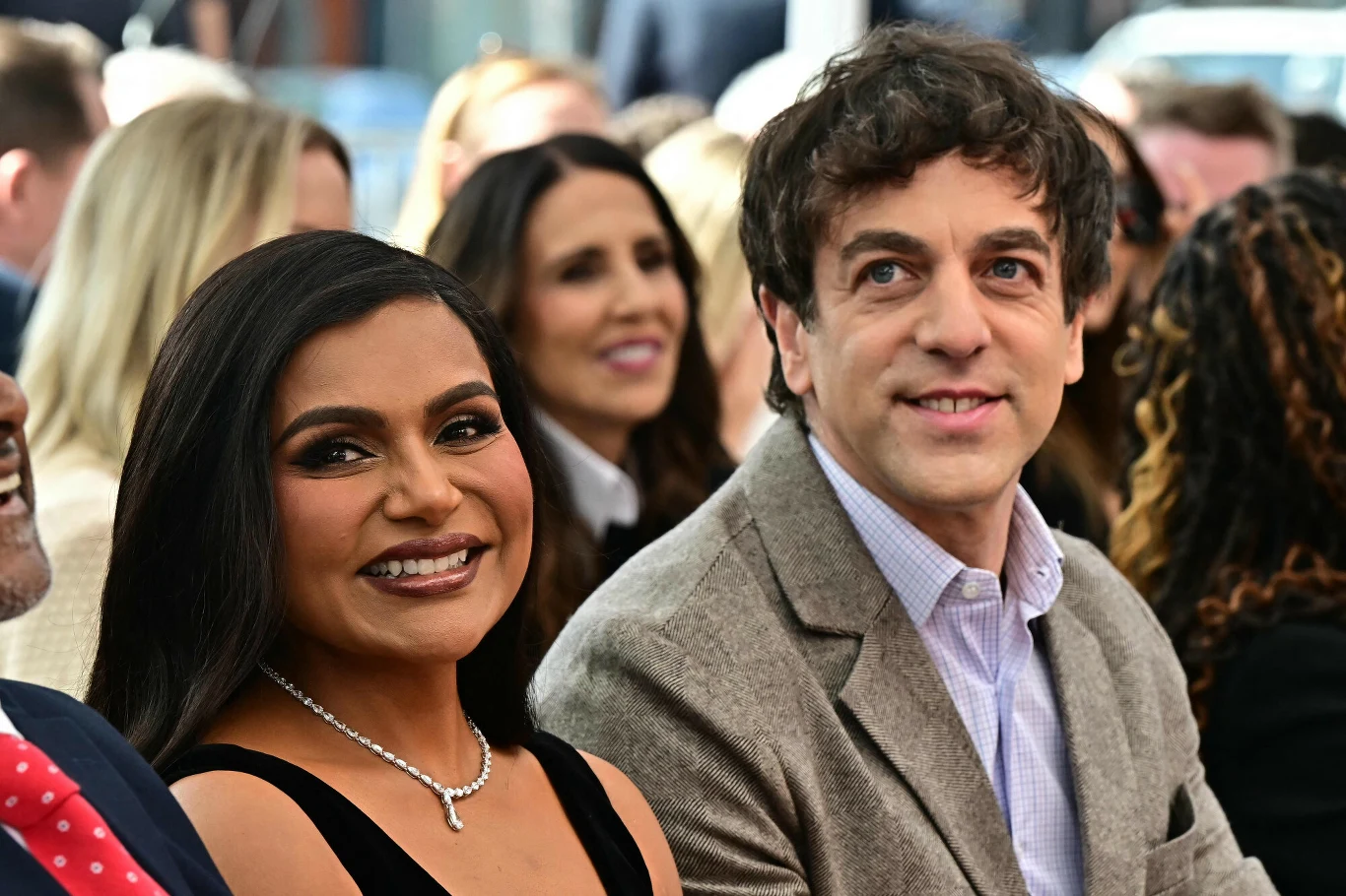 Mindy Kaling i B.J. Novak Uśmiechnięta kobieta w czarnej sukni i naszyjniku siedzi obok mężczyzny w szarej marynarce i jasnoniebieskiej koszuli wśród tłumu eleganckich osób.
