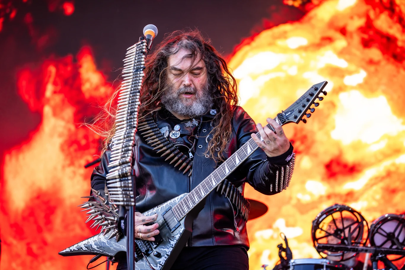 Max Cavalera stoi na czele grupy Soulfly Max Cavalera gra na gitarze elektrycznej, nosi nabojowy pas, w tle płonie kula ognia.