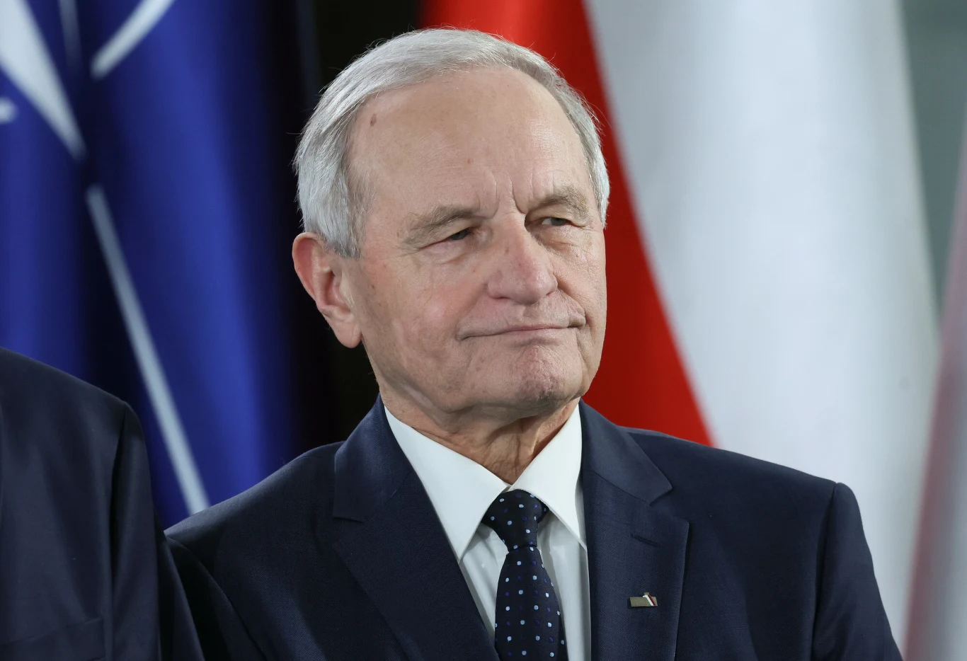 Były szef Biura Bezpieczeństwa Narodowego gen. Stanisław Koziej Starszy mężczyzna w garniturze i krawacie, stojący na tle flag w barwach biało-czerwonej oraz granatowej z białymi symbolami.