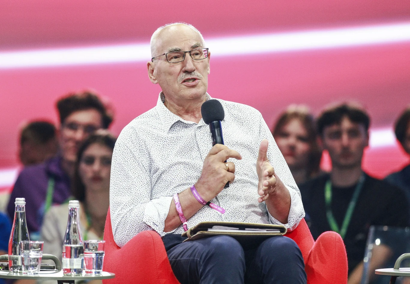 Starszy mężczyzna w okularach siedzi w czerwonym fotelu, trzyma mikrofon i gestykuluje podczas wypowiedzi, na kolanach ma notes; za nim znajduje się grupa słuchaczy.