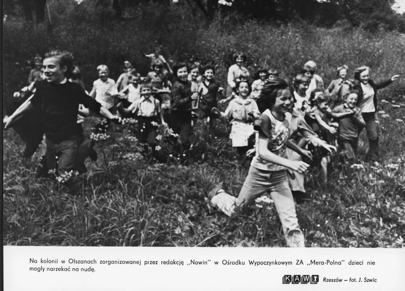 Grupa roześmianych dzieci biegnie przez łąkę, otoczona wysoką trawą i kwiatami, w pogodnej i beztroskiej atmosferze zabawy na świeżym powietrzu.