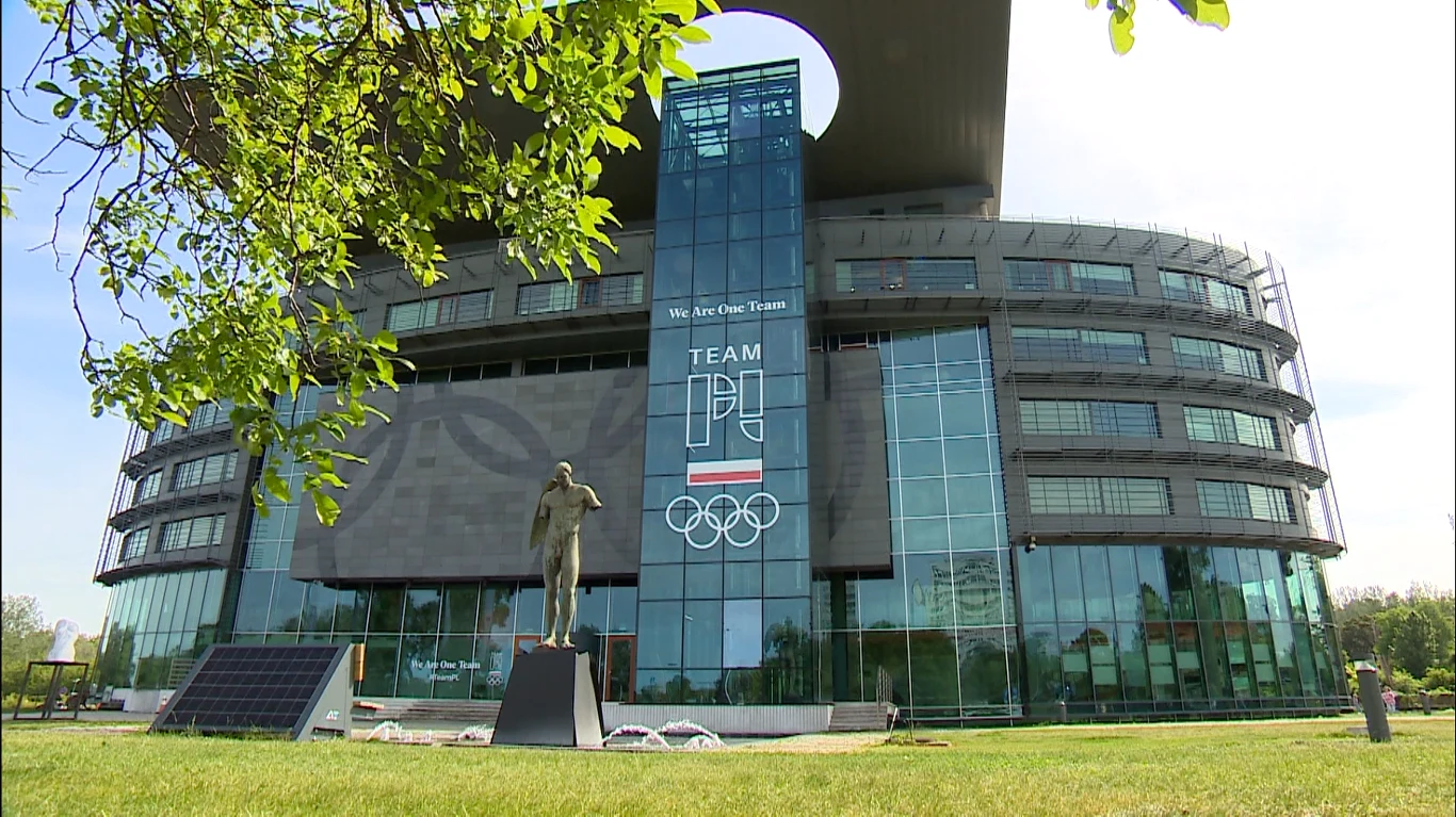 Nowoczesny budynek z przeszkloną fasadą, przed którym stoi pomnik sportowca na tle symboli olimpijskich i napisu TEAM POLSKA.