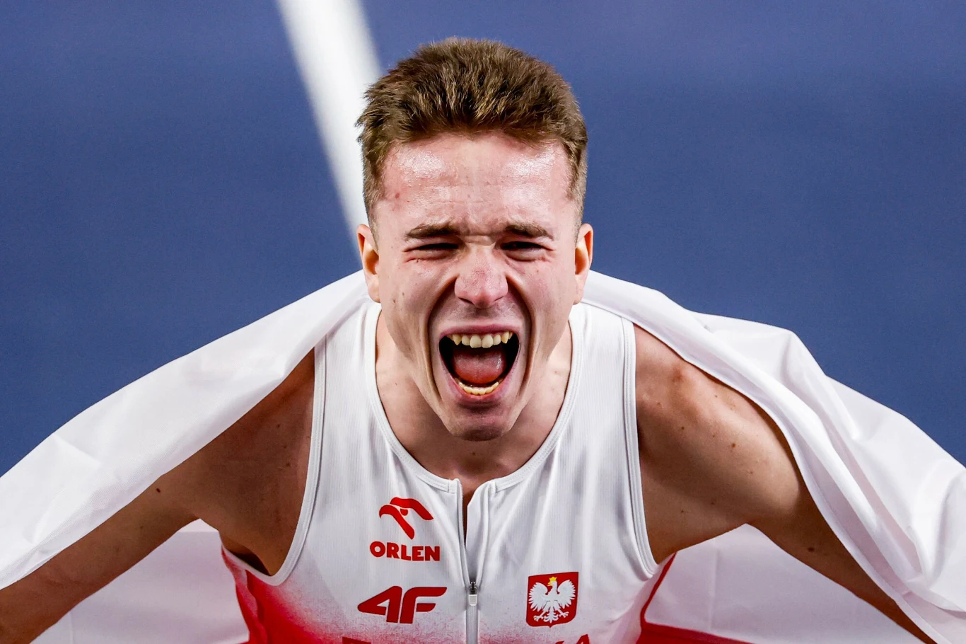 Mężczyzna w sportowym stroju z polskimi symbolami, z szeroko otwartymi ustami wyrażającymi silne emocje, owinięty biało-czerwoną flagą.