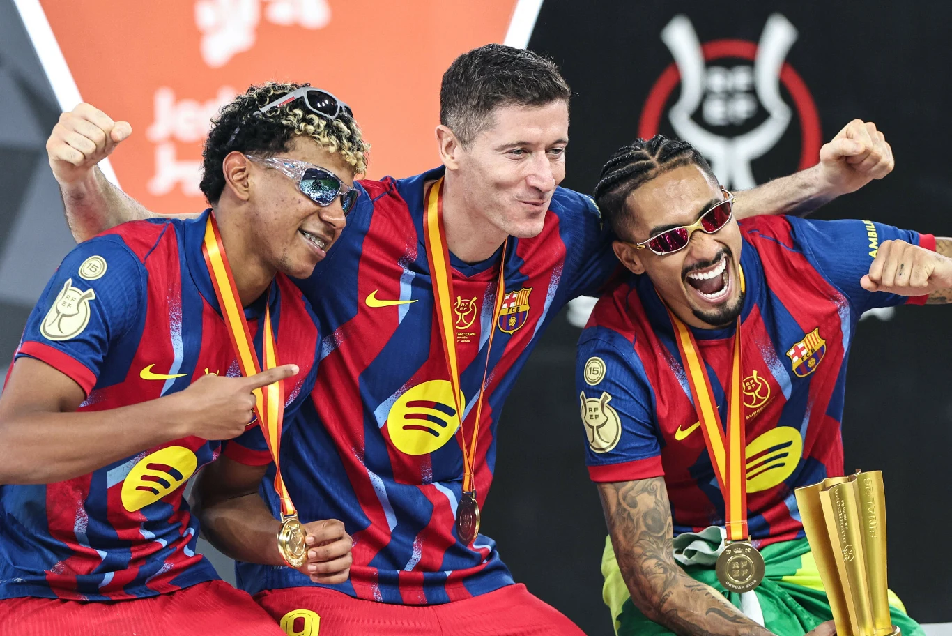 Trzech piłkarzy FC Barcelony w koszulkach klubowych świętuje z medalami na szyi, środkowy zawodnik obejmuje pozostałych, jeden z nich trzyma w ręku złoty puchar.