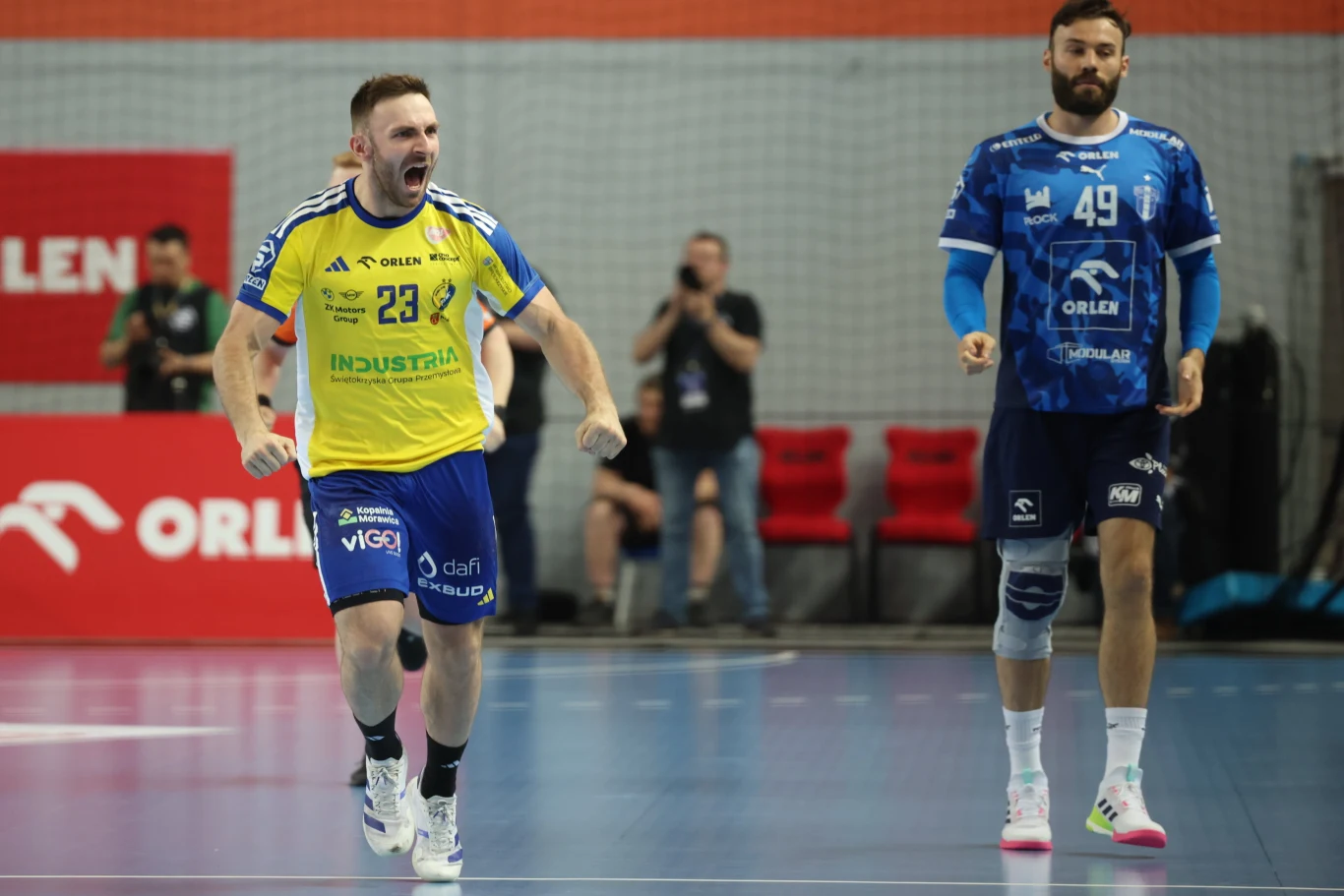 Zawodnik w żółto-niebieskim stroju energicznie celebruje zdobyty punkt na boisku do piłki ręcznej, obok niego stoi zawodnik drużyny przeciwnej w niebieskim stroju.