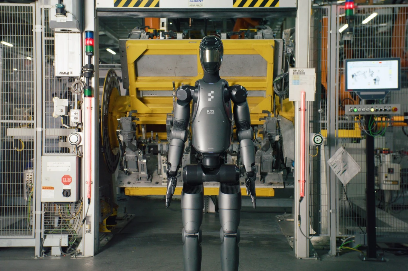 Humanoidalny robot stojący frontem w hali przemysłowej na tle dużej żółtej maszyny produkcyjnej i metalowych konstrukcji.