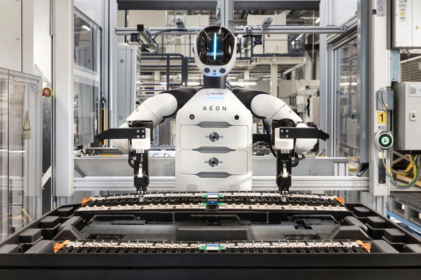 Humanoidalny robot z dwoma mechanicznymi ramionami wykonuje operacje montażowe przy dużym, przemysłowym urządzeniu elektrycznym w nowoczesnej hali produkcyjnej.