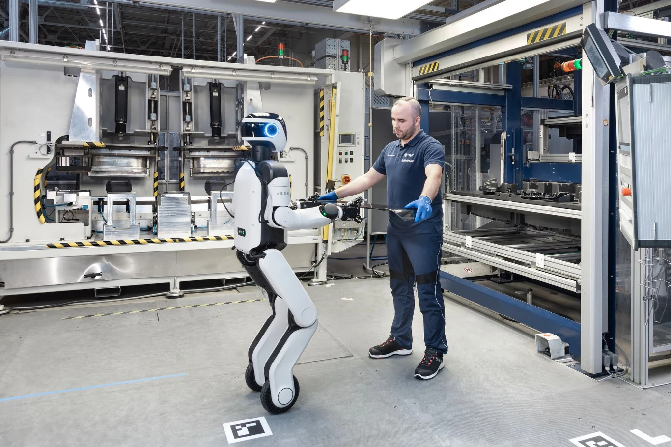 Człowiek ubrany w niebieski strój roboczy stojący naprzeciw humanoidalnego robota, wyciągającego do niego ręce w nowoczesnej hali produkcyjnej.