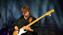 Mężczyzna grający na gitarze basowej podczas koncertu, skupiony na grze, ubrany w czarną koszulę, w tle zamglone światło sceniczne.