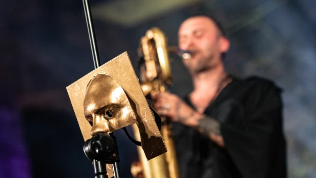 Metalowa maska w kolorze złota umieszczona na statywie na pierwszym planie, w tle mężczyzna grający na dużym instrumencie dętym, częściowo rozmyty, ubrany na czarno.