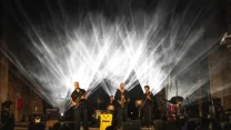 Zespół muzyczny grający na scenie, podkreślony efektownym oświetleniem tworzącym jasne snopy światła w tle.