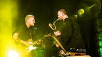 Dwóch muzyków na scenie: jeden gra na gitarze elektrycznej, drugi na saksofonie, obaj zwróceni ku sobie, oświetleni intensywnym żółto-zielonym światłem, z przodu widoczny fragment stolika z elektronicznym sprzętem.