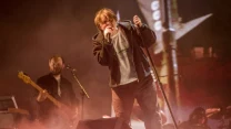 Wokalista trzyma mikrofon i śpiewa na scenie, w tle basista gra na gitarze oraz widoczni są pozostali członkowie zespołu przy instrumentach muzycznych, całość otulona światłem i mgłą sceniczną.