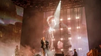 Wokalista wykonuje utwór na scenie przy intensywnym oświetleniu i wielkim ekranie z jego powiększonym wizerunkiem, a za nim zespół muzyczny gra na instrumentach klawiszowych i gitarze.