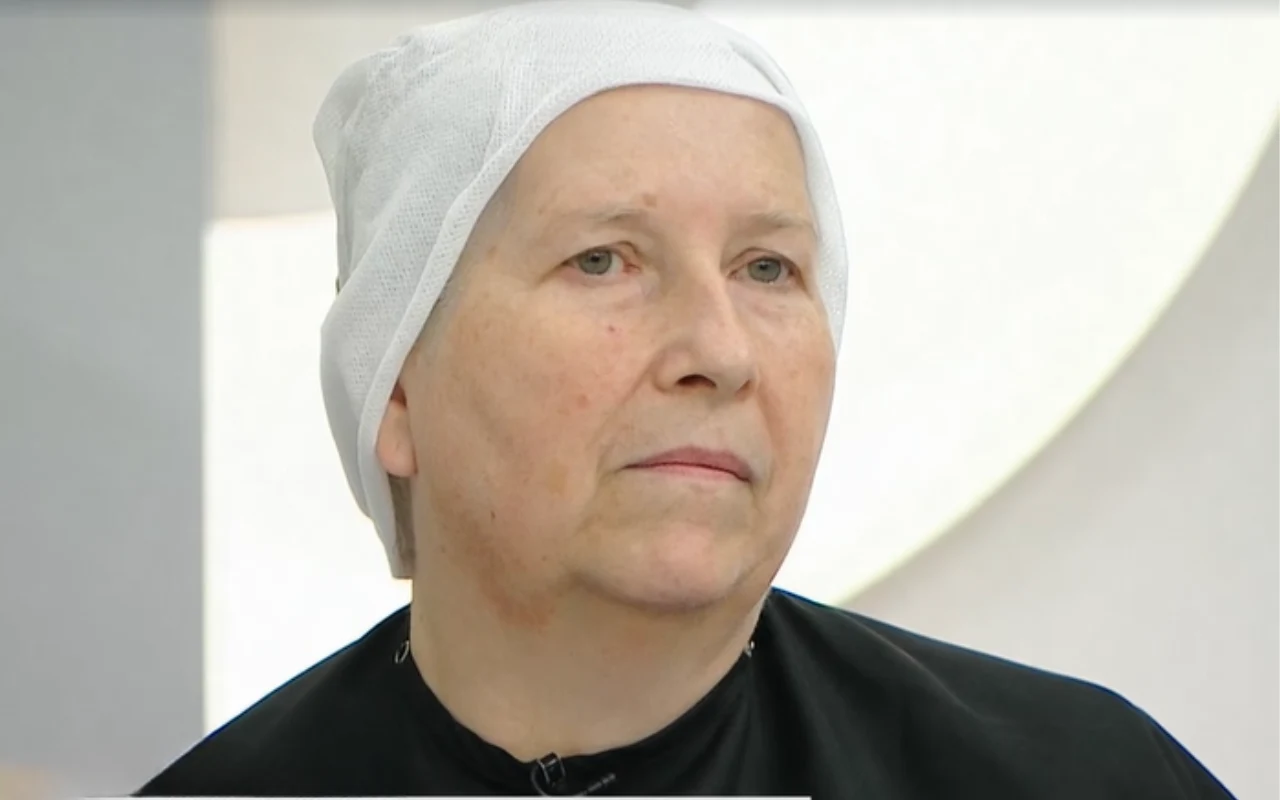 Dojrzała kobieta z lekko zarumienioną cerą, ubrana w czarną pelerynę fryzjerską i biały czepek ochronny na głowie, patrzy przed siebie w neutralnym wyrazie twarzy.