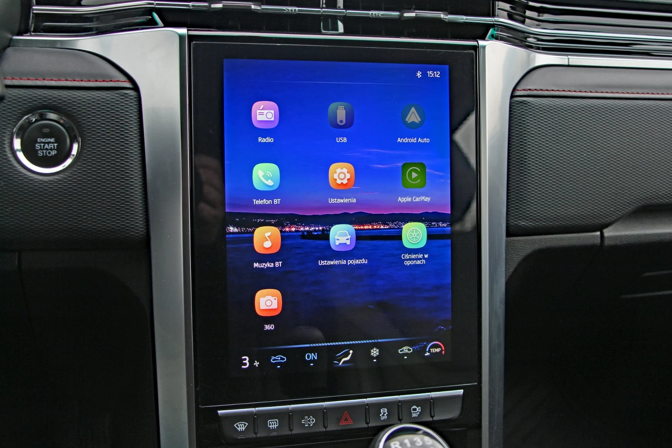 Duży ekran dotykowy systemu multimedialnego w samochodzie, z wyświetlonymi ikonami najważniejszych aplikacji, takich jak nawigacja, radio, USB, Android Auto i Apple CarPlay oraz przyciskami sterowania klimatyzacją pod ekranem.