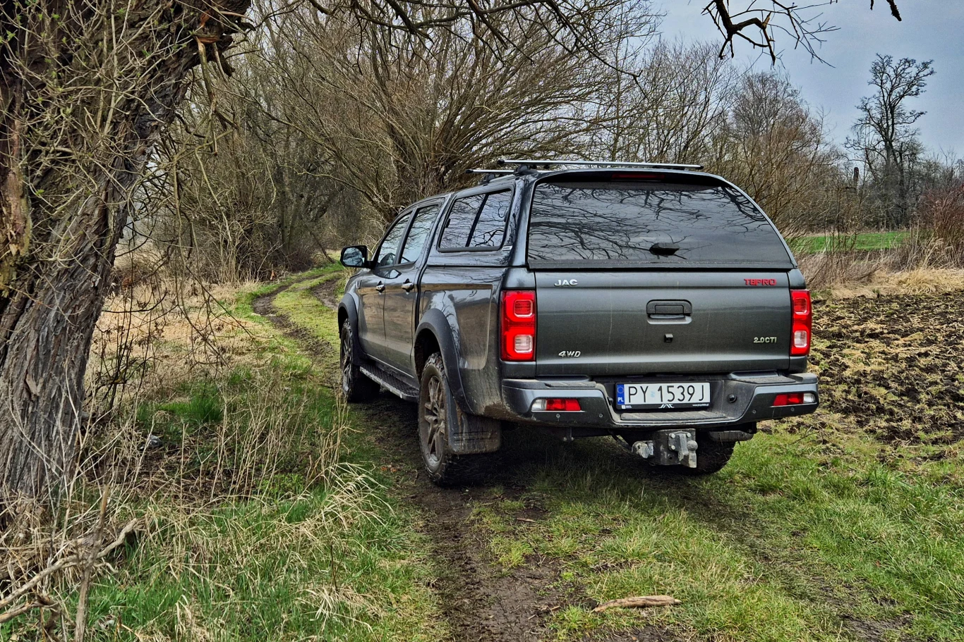 Szary pickup z zadaszoną paką porusza się wiejską, błotnistą drogą wśród drzew i traw.