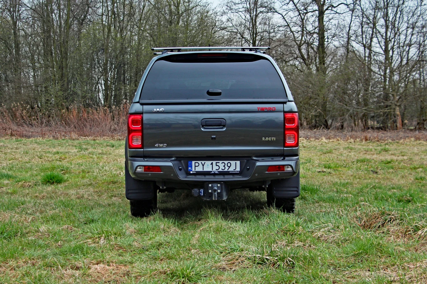 Szary pickup JAC T8 z zamkniętą paką, stojący na trawiastym polu na tle drzew.