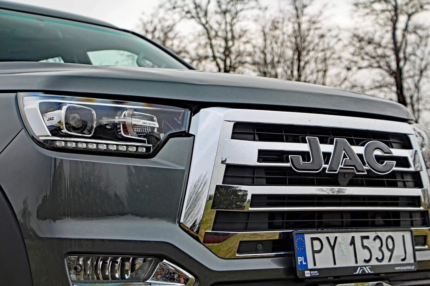 Duży chromowany grill z logo marki JAC, nowoczesny reflektor oraz tablica rejestracyjna pojazdu na tle rozmytych drzew bez liści.