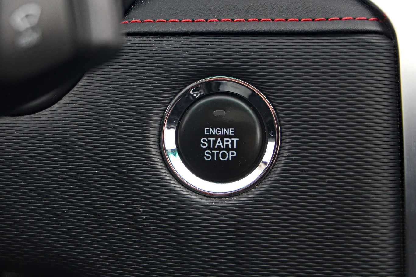 Przycisk uruchamiania i zatrzymywania silnika samochodu z napisem „ENGINE START STOP” otoczony srebrnym pierścieniem na desce rozdzielczej wykończonej czarną teksturą i czerwonym przeszyciem.