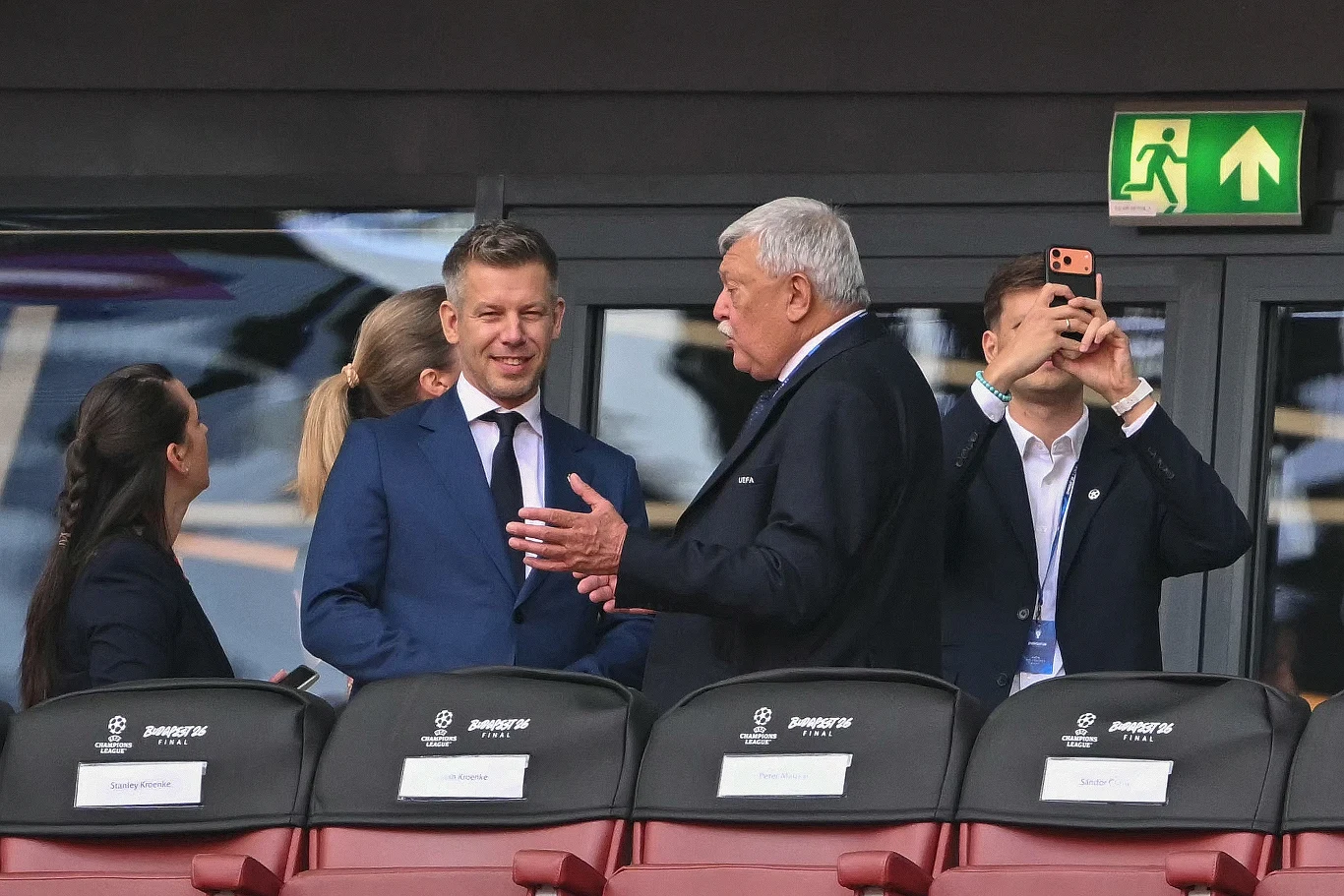 Dwóch elegancko ubranych mężczyzn w garniturach prowadzi rozmowę na trybunach stadionu, w tle osoba robi zdjęcie telefonem, a obok stoją dwie kobiety ubrane biznesowo.