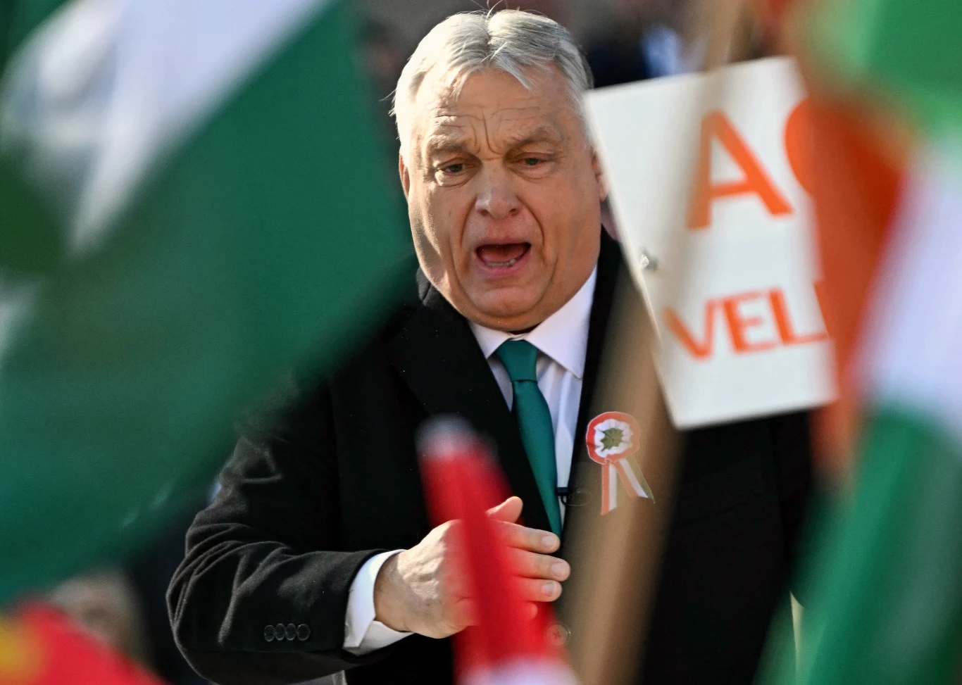 Były premier Węgier - Viktor Orban. Prywatnie wielki fan sportu Mężczyzna w garniturze z przypiętą rozetą i zielonym krawatem przemawia podczas wydarzenia, w otoczeniu flag i transparentów.