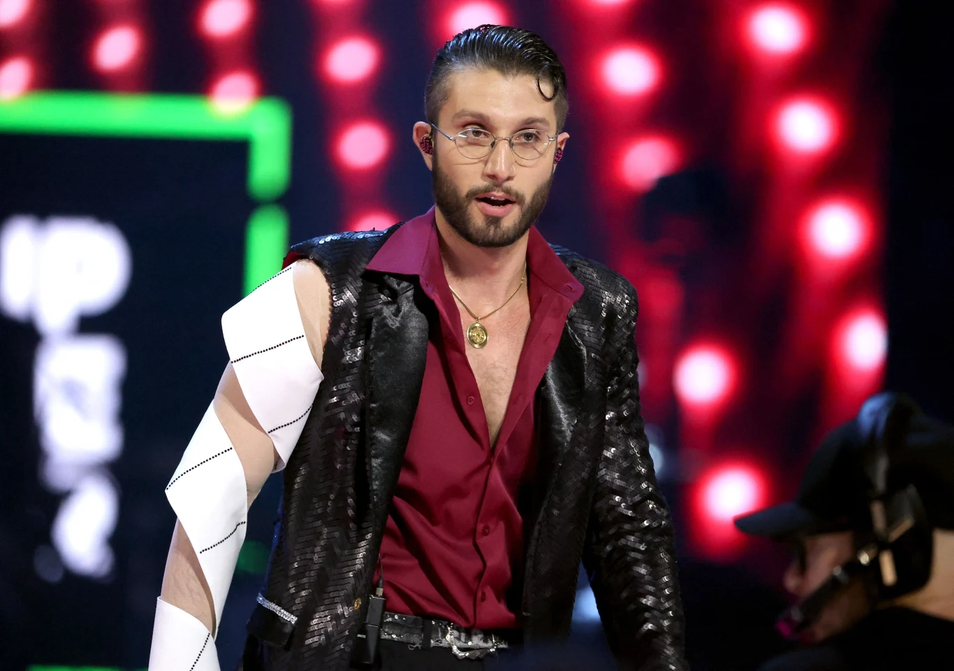 Mężczyzna w okularach i zarzuconym na ramieniu opatrunkiem, ubrany w połyskliwą czarną marynarkę i bordową koszulę, stoi na scenie oświetlonej czerwonymi światłami, w tle osoba z zestawem słuchawkowym.